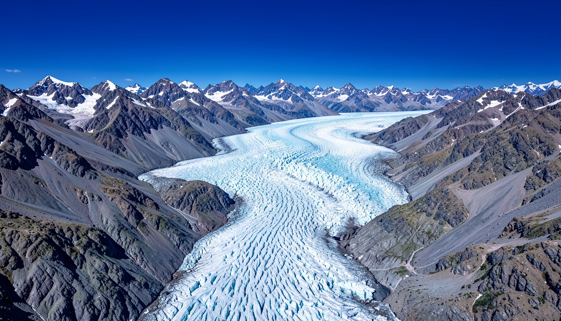Aerial view of a vast blue-white glacier winding through rugged mountain peaks under a clear sky