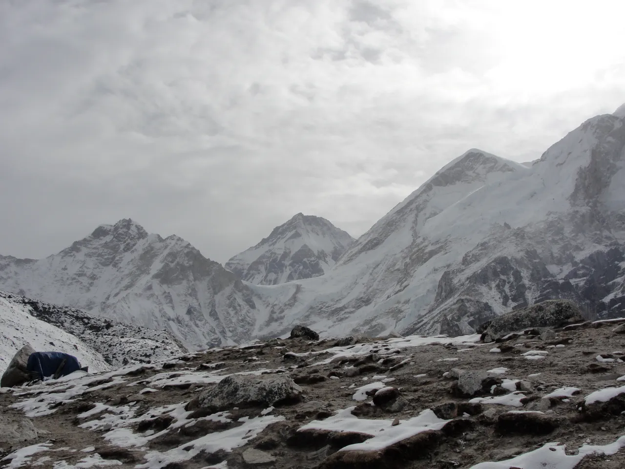 Wide view of snow-capped mountain peaks under a cloudy sky with rocky foreground, grey and white tones, dramatic mood