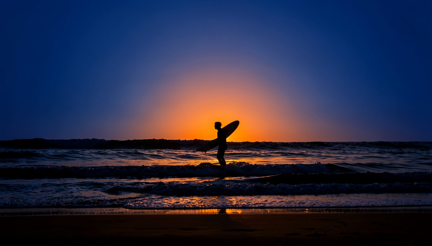 Surfer silhouetted against a warm orange sunset over gentle waves at a sandy beach