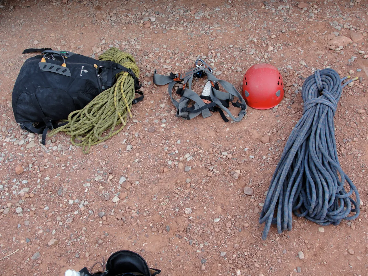 Climbing gear including coiled ropes, a backpack, harness, and red helmet laid out on reddish gravel ground, earthy tones
