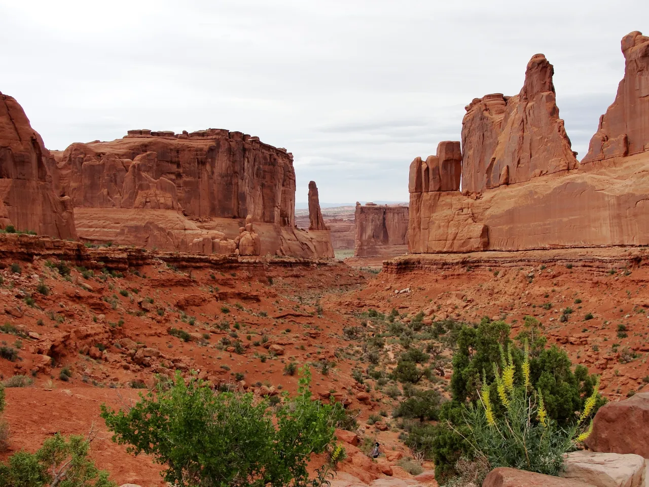 Desert landscape with tall red sandstone rock formations and green shrubs in foreground under cloudy sky, earthy mood