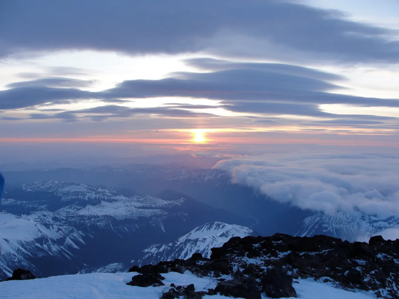 View from a high snowy mountain summit at sunset, layered clouds and distant ranges, cool blue and orange tones, dramatic