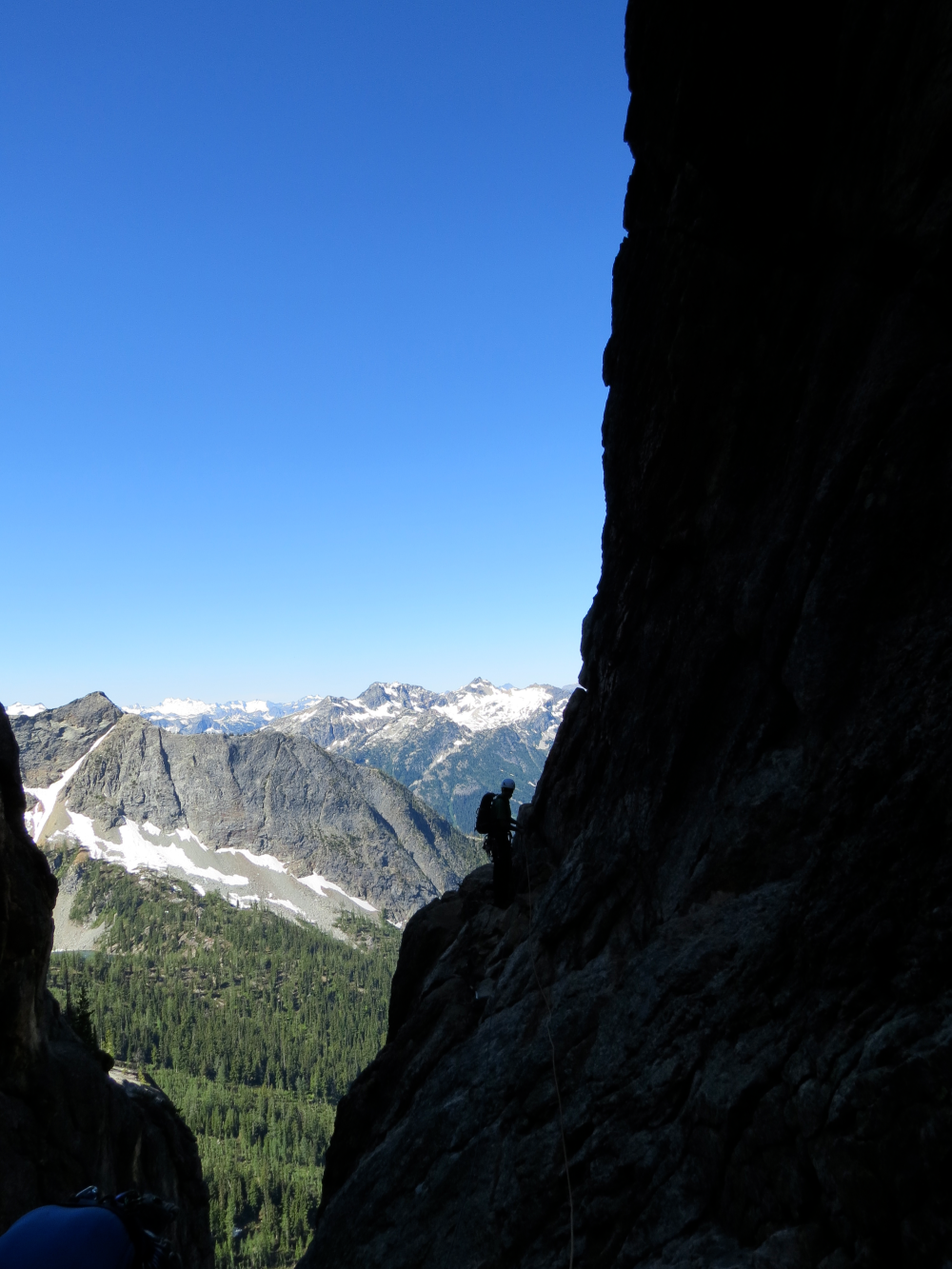 Silhouetted climber on a rocky cliff edge with expansive mountain range and forest valley in the background under clear sky
