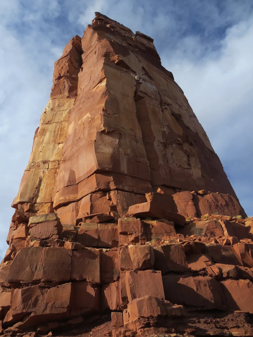 Tall reddish sandstone rock tower viewed from below against a blue sky with clouds